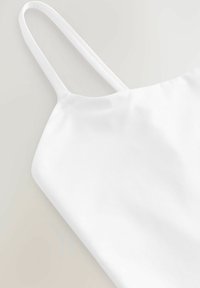 Valkoinen kapeahihnainen toppi, valmistettu sileästä kankaasta. Siisti pääntie ja pehmeästi laskeutuva muotoilu. Ei näkyviä kuvioita.