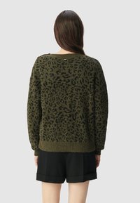 Vrouw met lang bruin haar, gekleed in een olijfgroen en zwart panterprinttrui en zwarte shorts met omgerolde pijpen, met haar rug naar de camera op een effen achtergrond.