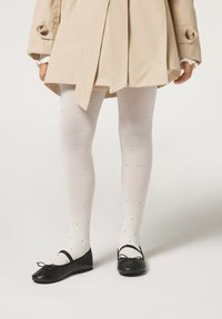 Beige Trenchcoat, weiße gemusterte Strumpfhosen mit goldenen Punkten und schwarze Ballerinas mit Schleifen. Merkmale sind eine glatte Stofftextur und ein schlichtes Design.