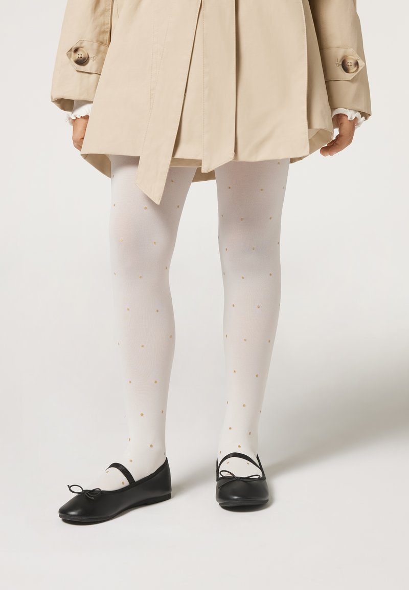 Beige Trenchcoat, weiße gemusterte Strumpfhosen mit goldenen Punkten und schwarze Ballerinas mit Schleifen. Merkmale sind eine glatte Stofftextur und ein schlichtes Design.