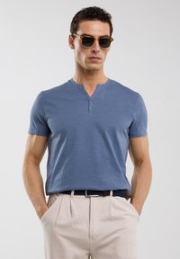 Modrá tričko s krátkým rukávem henley s kulatým výstřihem a lištou na tři knoflíky, nošeno s světlými kalhotami a slunečními brýlemi.