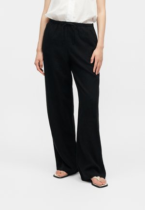 Žena nosí čierne široké nohavice s elastickým pásom a biely bezrukávový top s gombíkmi, skombinované so sandálmi so štvorcovou špičkou.