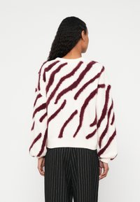 Nő fehér pulóverben, amelyen bolyhos, átlós bordó csíkok vannak, és fekete, vékony csíkos nadrágot visel, hátulnézetben, egyszerű háttér előtt.