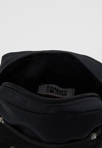 Borsa nera in nylon con superficie testurizzata, dotata di un compartimento principale e di una tasca con zip, etichettata con "TOMMY JEANS" all'interno.