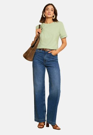 Kobieta ubrana w jasnozielony krótki rękaw, niebieskie szerokie dżinsy, brązowe sandały na obcasie, duże kolczyki koła, trzyma brązową zamszową torebkę na ramię.