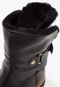 Bota de tobillo de cuero negro con un suave forro de piel negra, un acento de botón dorado y un exterior liso con detalles de costura curva.