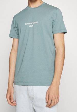 Türkisfarbenes Baumwoll-T-Shirt mit Rundhalsausschnitt, kurzen Ärmeln und einem weißen, aufgedruckten Logo mit der Aufschrift "EMPORIO ARMANI MILANO" auf der Brust.