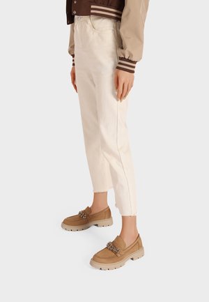 Mocassini marroni con suole spesse, adornati con accessori decorativi. Indossati con pantaloni cropped color crema con orli sfrangiati.
