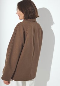 Brūns vilnas maisījuma mēteles ar brīvu piegriezumu, augstu apkakli un redzamām šuvēm. Garās piedurknes un smalkā tekstūra papildina tās dizainu.