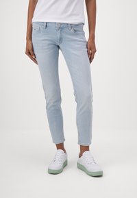 Jeans de corte entallado en azul claro, con cintura de altura media, bolsillos frontales y un desgaste desgastado, combinados con zapatillas blancas con detalles en verde.