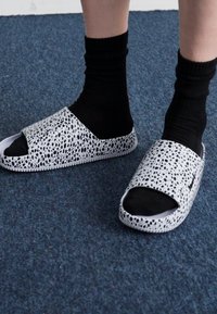 Sandálias slide de dedo abertas, com um design manchado em preto e branco, apresentando um cabedal suave e texturizado e uma sola robusta. Usadas com meias pretas.