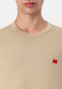 Beige gestrickter Pullover mit rundem Ausschnitt, ausgestattet mit einem kleinen roten Logo-Patch. Die Textur wirkt glatt und leicht.