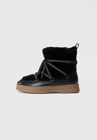 Botte d'hiver noire avec doublure en fourrure, lacets enroulés, détails en cuir et semelle en caoutchouc marron épaisse, vue de côté.