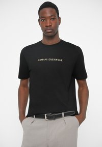 Μαύρο κοντομάνικο πουκάμισο με "ARMANI EXCHANGE" σε χρυσά γράμματα, φορεμένο από έναν άνδρα με τα χέρια στις τσέπες, ανοιχτό γκρι παντελόνι και μαύρη ζώνη.