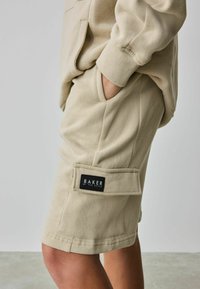 Pantalones cortos de algodón beige con un bolsillo lateral, que presentan una etiqueta negra que dice "BAKER by Ted Baker". Textura suave con sutiles detalles de costura.