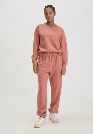 Rosa Baumwollhoodie und passende Jogginghosen mit einem Kordelzugbund. Texturiertes Material, lockere Passform, kombiniert mit weißen Turnschuhen.