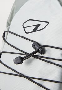 Lys grå ryggsekk i materiale med teksturert overflate, med svarte bungee-korder og en fremtredende svart logo. Justerbare hardwaredetaljer er synlige.
