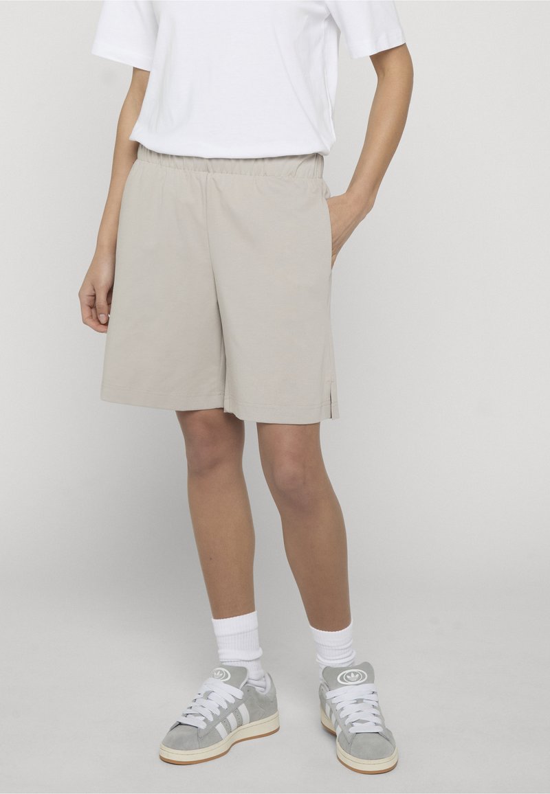 Personne portant un short beige à taille élastique, un t-shirt blanc, des chaussettes blanches montantes et des baskets Adidas grises, debout avec une main dans la poche.