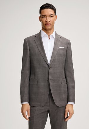 Man draagt een grijs geruit tweeknoops kostuumjasje met bijpassende broek en een wit overhemd tegen een effen achtergrond.