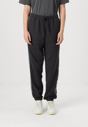 Pantalones de chándal negros con cintura elástica y cordón, que presentan coloridos patrones florales en los laterales y dobladillos ajustados. Llevados con zapatillas blancas.
