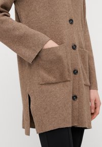 Hnědý pletený cardigan s knoflíkovým zapínáním, dvěma předními kapsami a bočními rozparky pro snadný pohyb. Texturovaná tkanina s jemným povrchem.