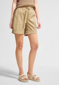 Khakinväriset shortsit, joissa on joustava vyötärönauha ja nyöri, kaksi etutaskua ja käännetyt lahkeensuut. Käytetty vaaleiden sandaalien kanssa.