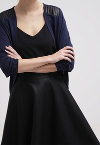 Námořnický svetr na knoflíky s žebrovanými koženými akcenty na ramenou, přes černý top s výstřihem do V, doplněný vysokým pasem černé kožené sukně.