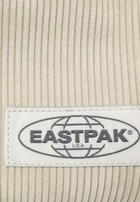 Béžová manšestrová tkanina s vertikálním ribbovaným vzorem. Bílá etiketa zobrazuje "EASTPAK" v šedé barvě, orámovaná grafickým symbolem zeměkoule.