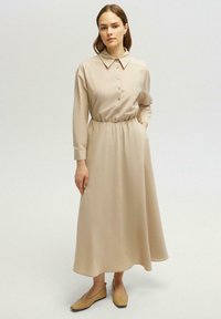Robe chemise beige à manches longues avec une fermeture à boutons, taille élastique et poches latérales. Portée avec des ballerines marron clair. Texture lisse.