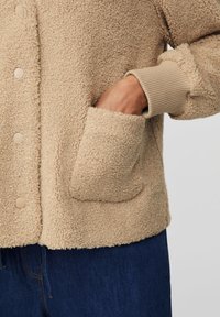 Beżowa, pluszowa, miękka kurtka o fakturowanej powierzchni, z prążkowanymi mankietami i dużymi bocznymi kieszeniami; zapinana na jasne guziki.