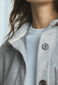 Grauer Wollmantel mit einem taillierten Kragen und einem hellblauen Kurzarmhemd darunter. Verfügt über eine Knopfleiste und ein Etikett entlang des Kragens.