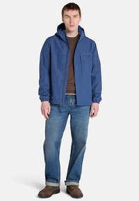 Blå vattentät jacka med huva, två framfickor och elastiska ärmslut, bärs över en brun skjorta och blå jeans. Bruna skor fulländar outfiten.