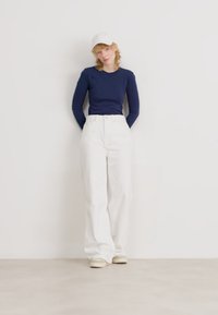 Camisa de manga larga azul marino, pantalones de pierna ancha blancos y zapatos beige combinados con una gorra blanca. El modelo está de pie contra un fondo claro.