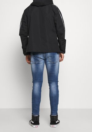 Zwarte hoodie jacket met reflecterende zijstrepen, gecombineerd met aansluitende blauwe spijkerbroek en zwarte hoge sneakers met witte details.