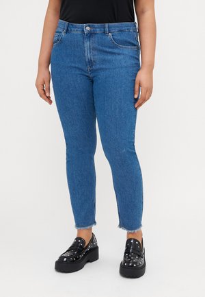 Blue denim jeans with a slim fit, featuring a mid-rise waist, subtle whiskering, and raw hem accents. Paired with black platform loafers.