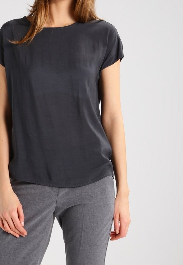 Femme portant un chemisier gris foncé à manches courtes et un pantalon gris clair, debout devant un fond clair uni.