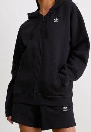 Osoba nosząca czarną bluzę Adidas z zamkiem błyskawicznym i przednimi kieszeniami oraz pasujące czarne spodenki Adidas z logo na lewej nodze.