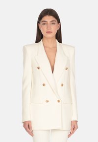 Blazer bianco doppiopetto con rever a punta, bottoni dorati e tasche a toppa. Texture liscia, vestibilità su misura e spalline per una maggiore struttura.