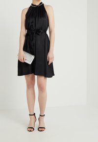 Robe noire à col arrondi et à bretelles, avec un col volant, une ceinture à la taille et une jupe fluide ; pochette argentée à la main, associée à des talons noirs à lanières.