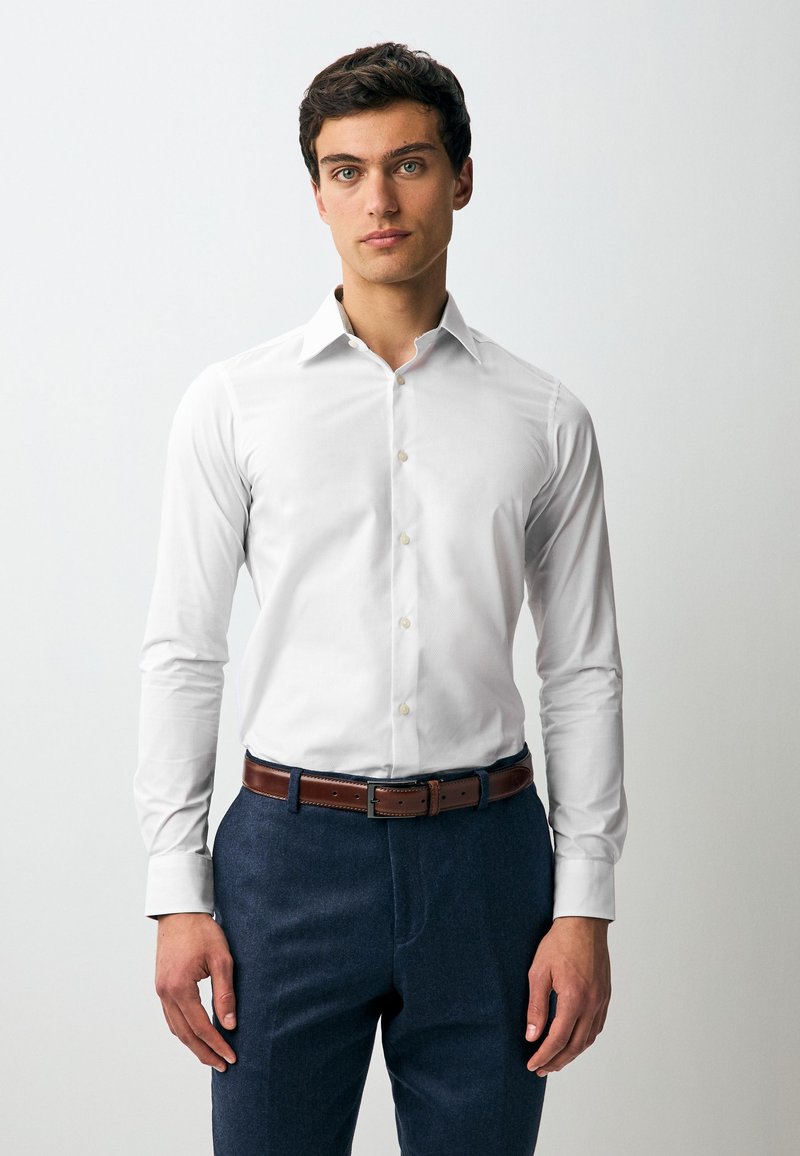 Camicia bianca a maniche lunghe con colletto appuntito, apertura frontale con bottoni e trama sottile. Indossata con pantaloni sartoriali blu scuro e una cintura marrone.