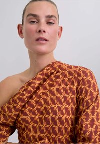 Patroon, asymmetrische top met een roestkleurige basis en een geometrisch gele print. Gemaakt van zachte stof met een gedrapeerd ontwerp.