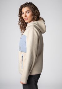 Vrouw met krullend haar die een crèmekleurige fleecetrui met een lichtblauw borstzakje draagt, glimlacht en over haar schouder kijkt tegen een effen achtergrond.