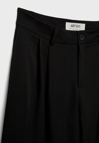 Zwarte high-waisted broek met een knoop en riemlussen, met plooien nabij de tailleband en een zichtbaar merkteken aan de binnenkant.