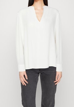 Vrouw draagt een wit blouse met lange mouwen en een V-hals, gecombineerd met een zwarte spijkerbroek, staand tegen een effen lichte achtergrond.