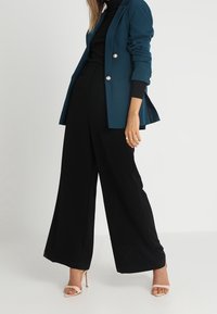 Zielony marynarka z srebrnymi guzikami, czarny top z wysokim kołnierzem oraz szerokie czarne spodnie. Noszone z beżowymi szpilkami. Gładka tekstura.