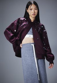 Chaqueta bomber burdeos con lentejuelas reflectantes, puños acanalados y cremallera. Llevada sobre un top corto gris claro y pantalones de chándal a juego.