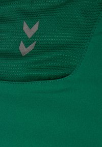 Grünes Sportoberteil mit einem strukturierten Muster, glattem Stoff und zwei grauen Chevron-Akzenten am Halsauschnitt.