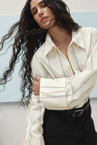 Kremo spalvos marškiniai su pilkomis juostelėmis, oversized rankogaliai, laisva forma, dėvimi su juodomis kelnėmis. Lygus audinys, standartinis apykaklė, mygtukų užsegimas.