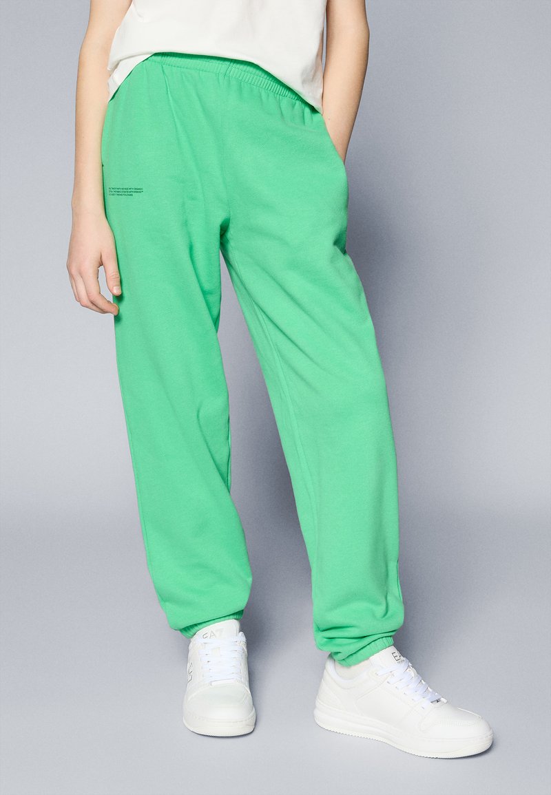 Pantalones de sudadera verdes con cinturilla elástica, bolsillos laterales y tobillos acanalados. Combinados con zapatillas blancas. La tela parece suave y ligera.