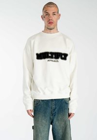 Weißes Sweatshirt mit dem Aufdruck "MULTIPLY APPAREL" in Schwarz. Verfügt über gerippte Bündchen und Saum, kombiniert mit lockeren blauen Jeans.
